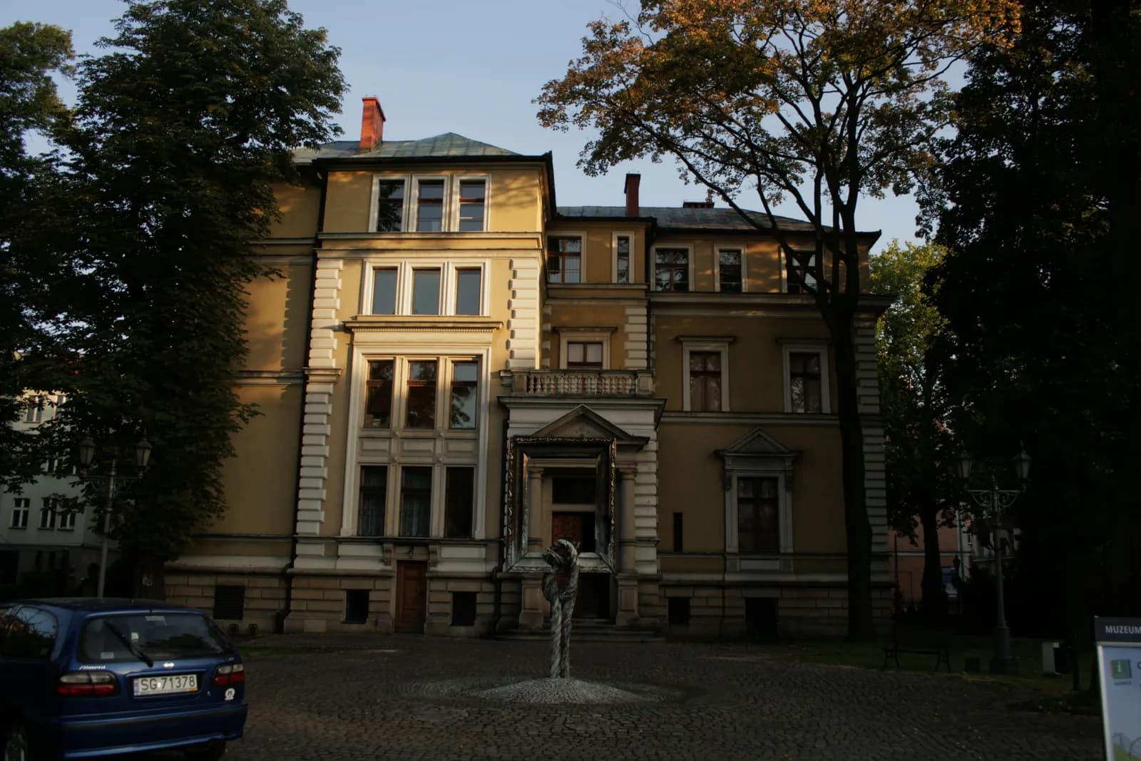 A grand, multi-story yellow building with many windows and a large entrance framed by trees, with a blue car parked on the left and a small sculpture in front.