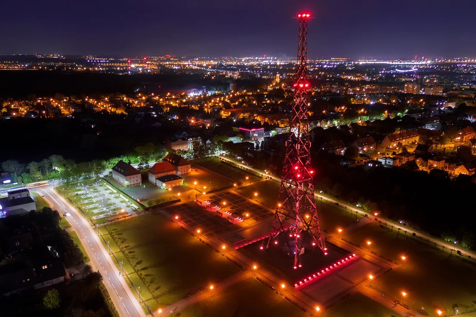 An aerial night view shows a tall radio tower illuminated with red lights, standing prominently against a brightly lit city skyline.
