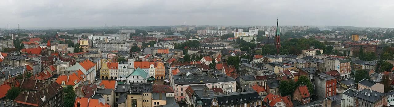 A wide, panoramic view of a city with a mixture of old and modern buildings, with a prominent green-spired church tower rising above the rooftops.